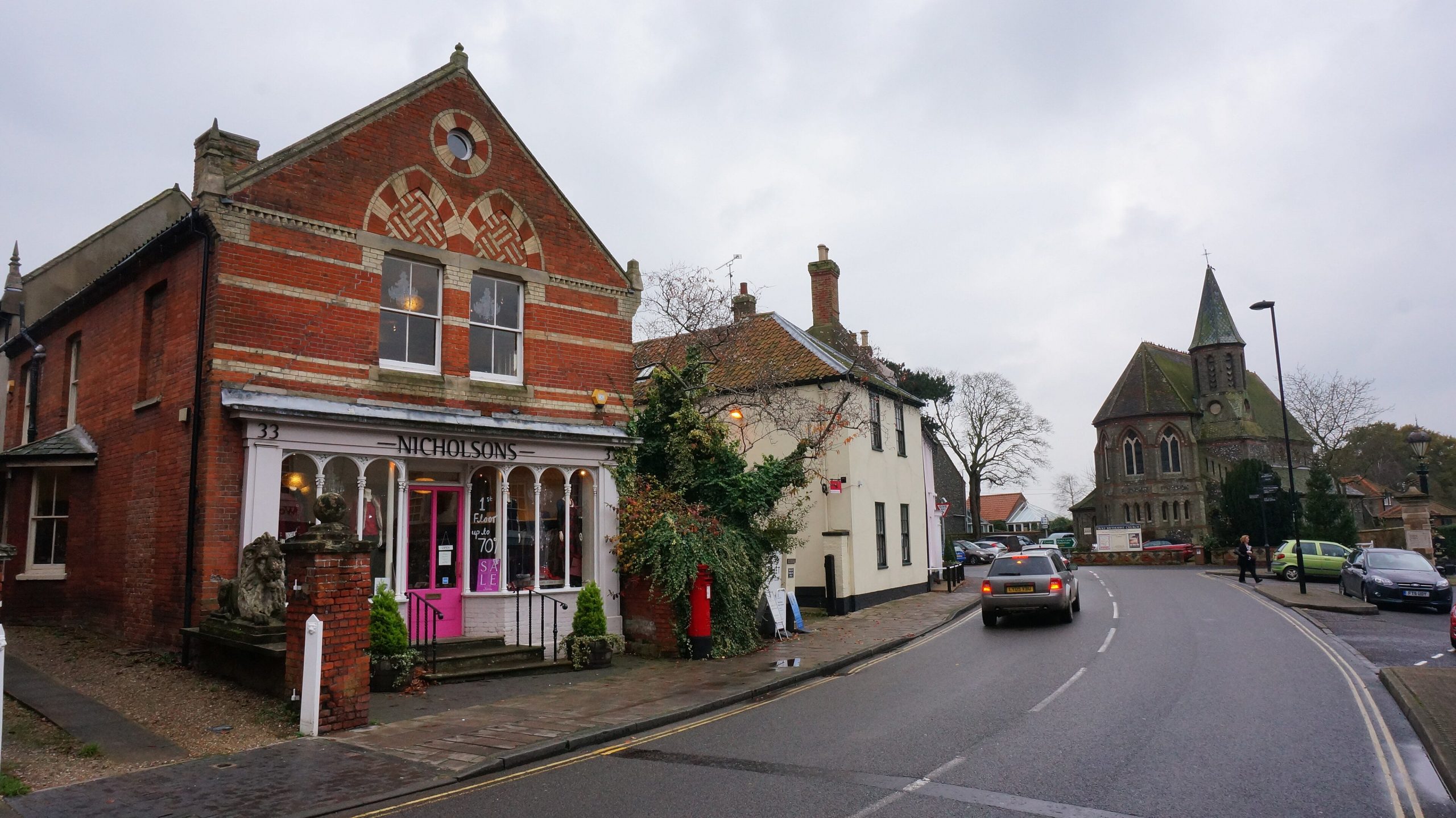 A red-brick shop on the High Street in Holt, with a church spire beyond